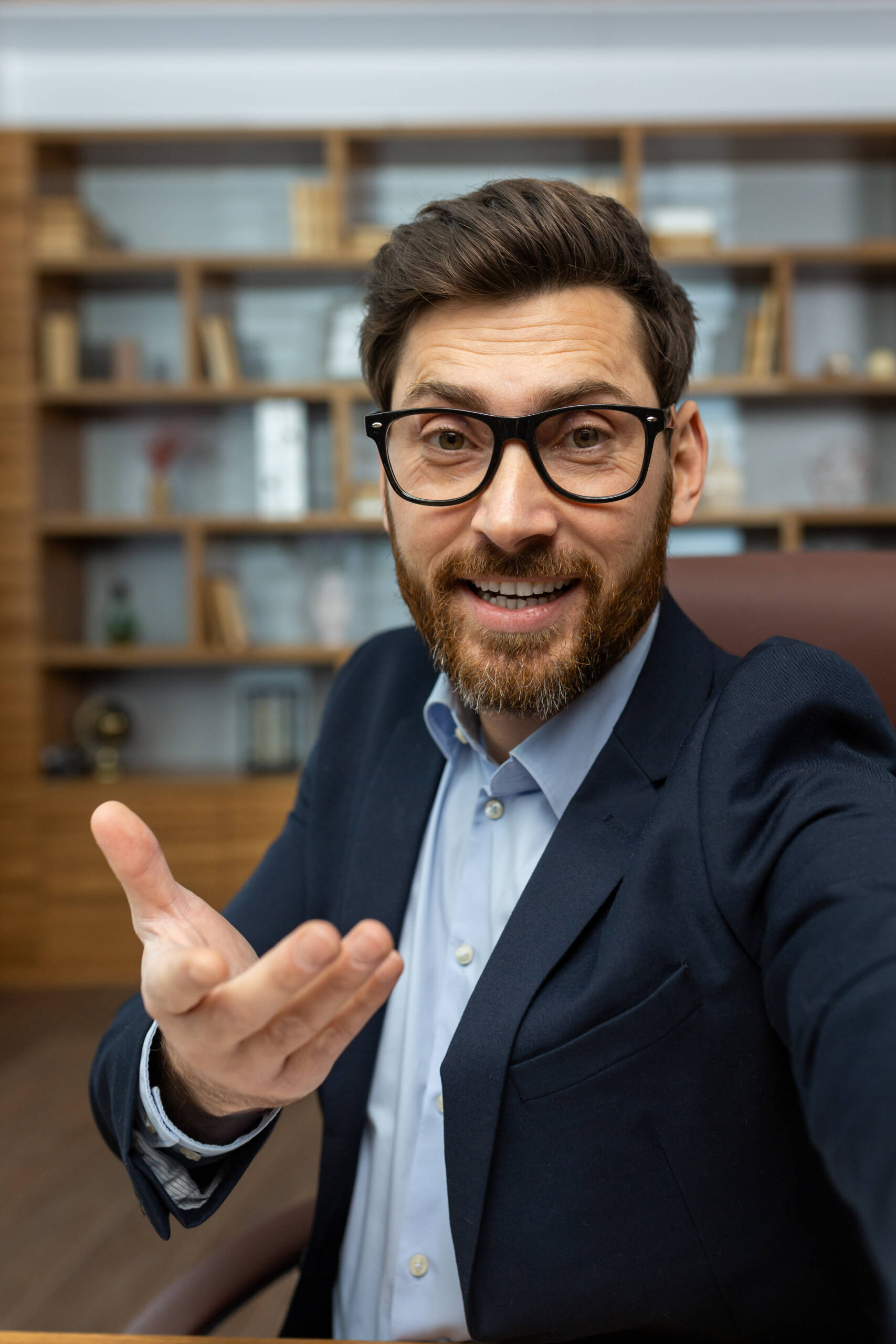 Vertical close-up photo of a young man in a business suit and glasses sitting in the office, talking online and gesturing to the camera of the device.
